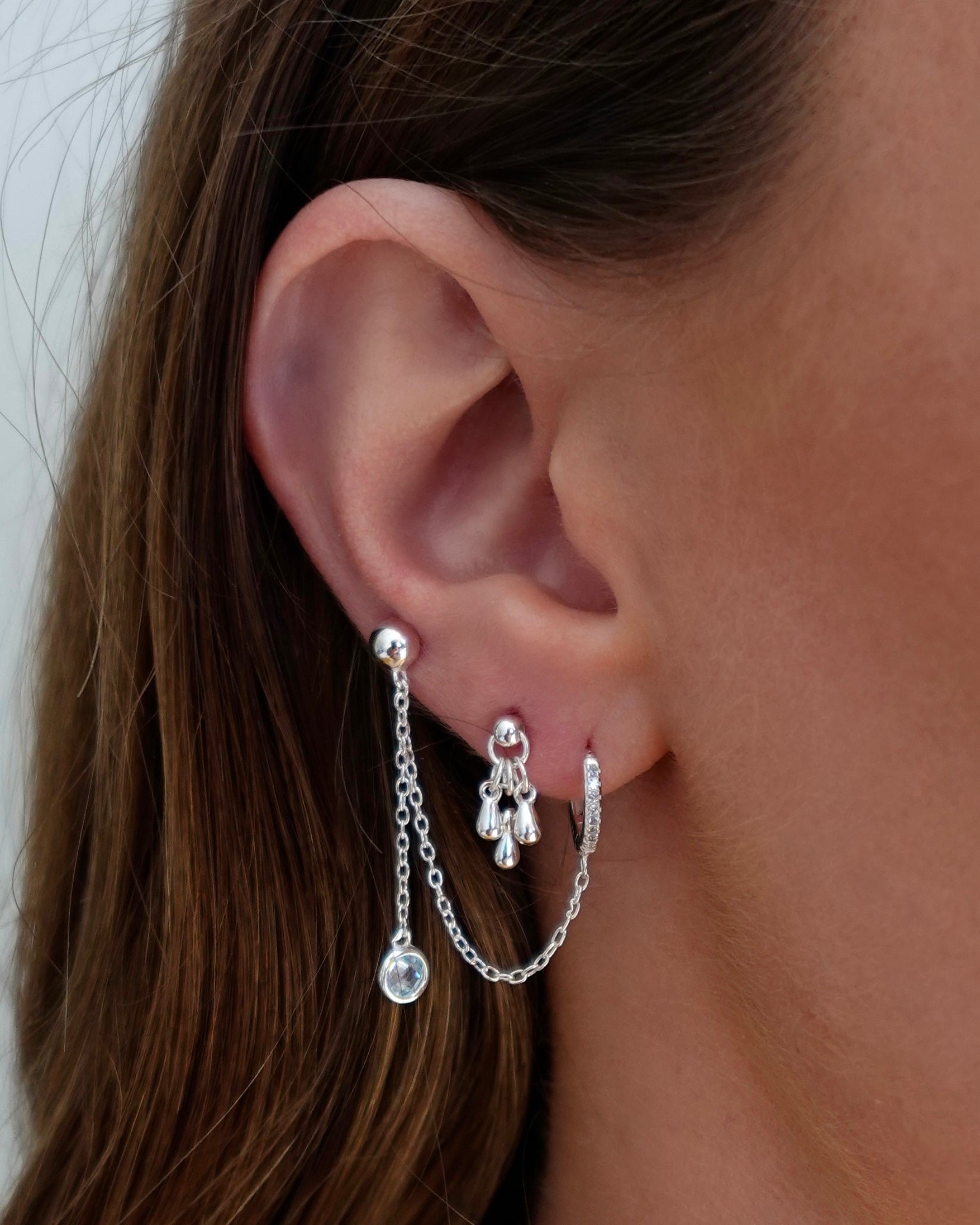 Close-up of an ear wearing silver hoop earrings with chain and crystal details.